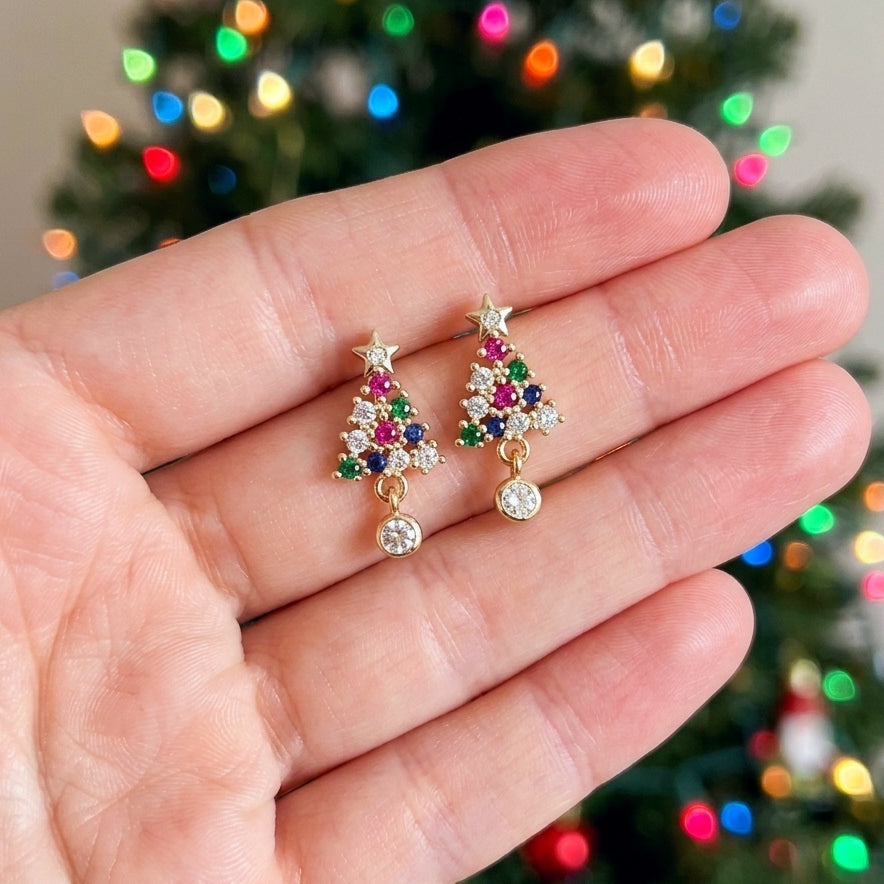 A pair of gold Christmas tree stud earrings with colorful gemstone accents, held in a person's hand with a blurred Christmas tree in the background.
