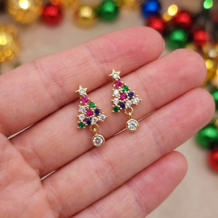A pair of gold Christmas tree stud earrings with colorful gemstone accents, held in a person's hand against a festive background of blurred holiday ornaments.