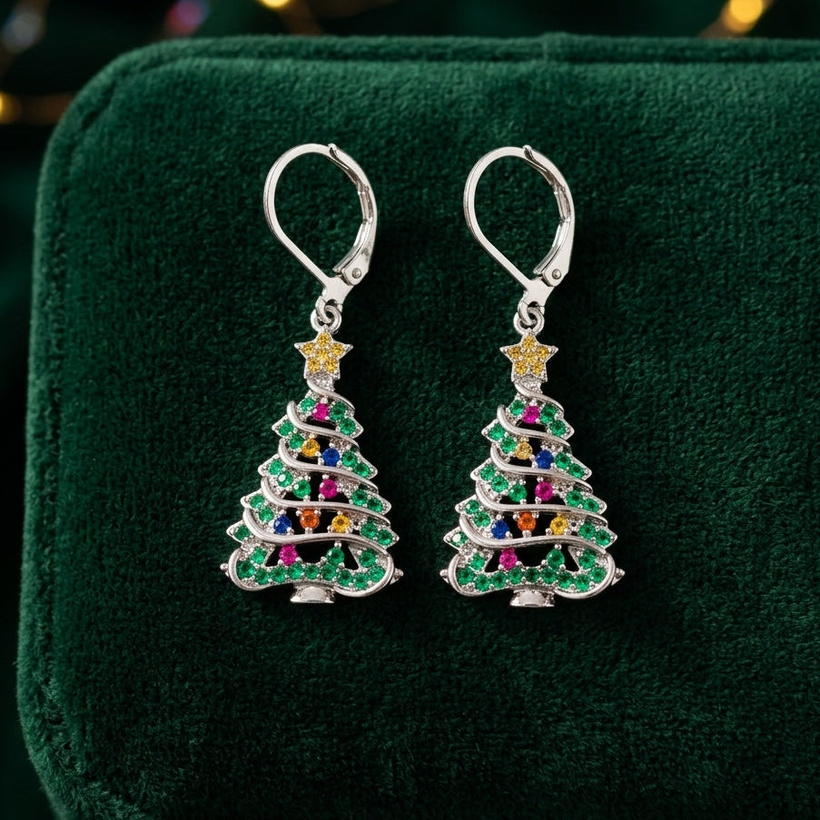 A pair of silver leverback earrings shaped like Christmas trees, decorated with green, pink, blue, yellow, and orange rhinestones, displayed on a dark green velvet jewelry box.