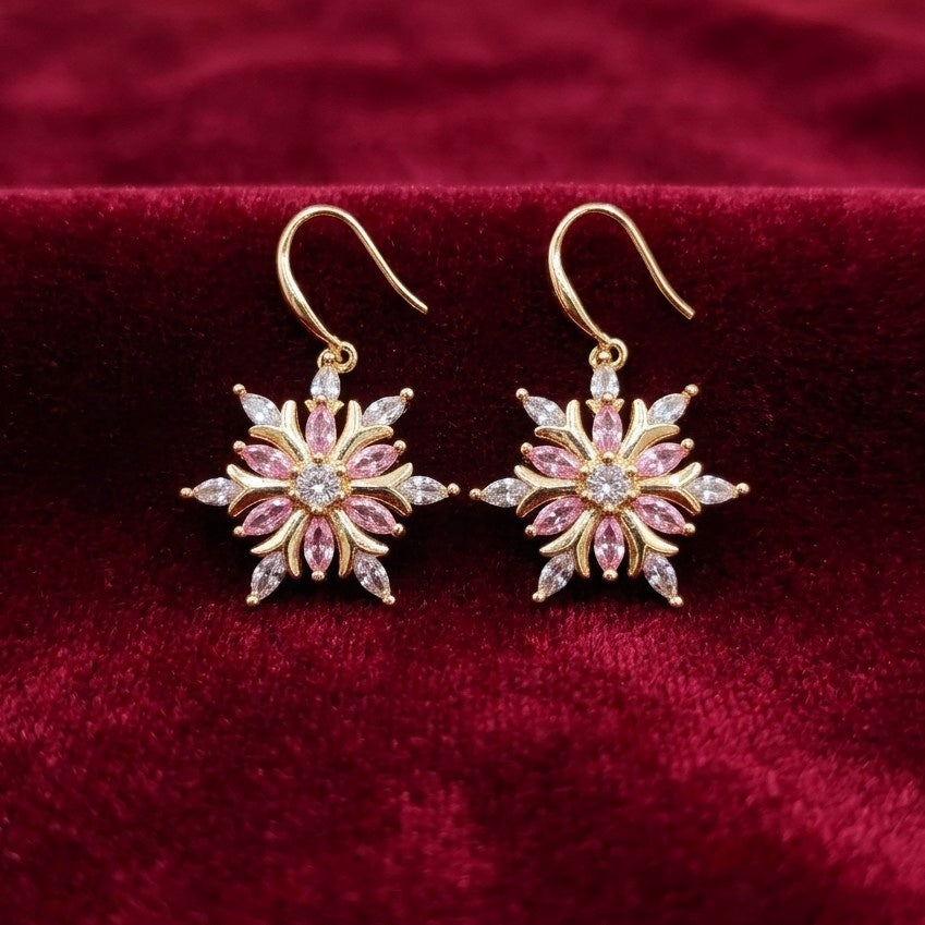 A pair of gold-tone snowflake dangle earrings with pink and clear cubic zirconia crystals, displayed on a deep red velvet surface.