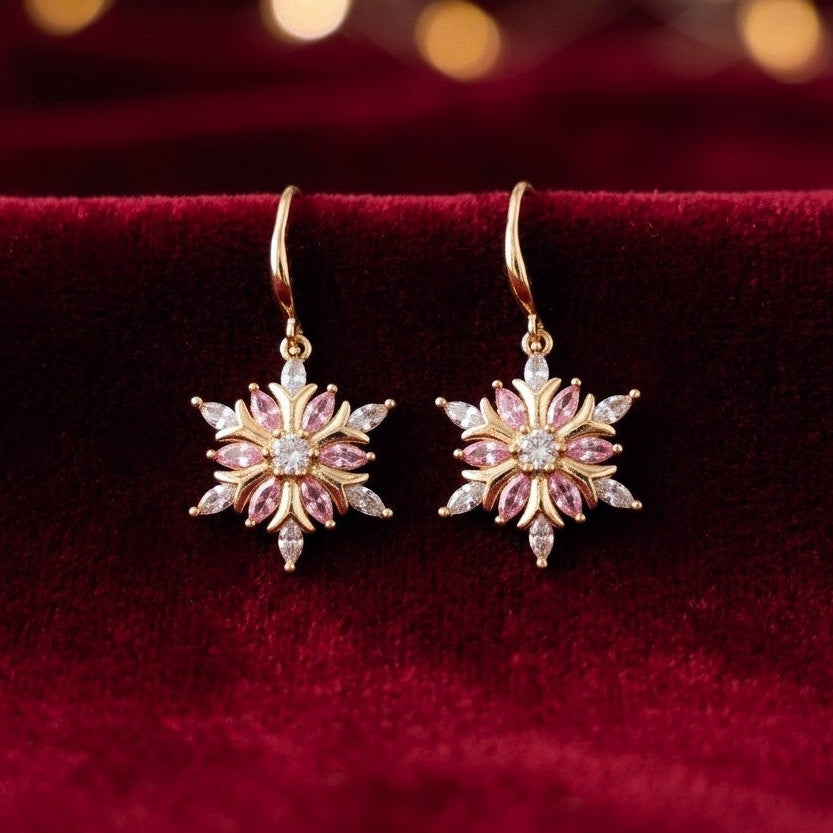 A pair of gold-toned dangle earrings with pink gemstone floral snowflake designs and clear center stones, displayed on a deep red velvet surface with a softly blurred warm background.