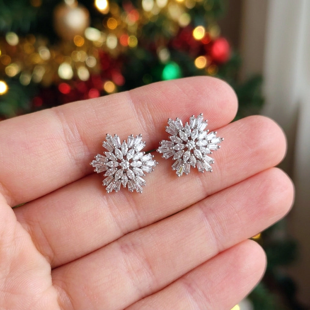A pair of silver snowflake stud earrings held on a person's fingers, with a blurred Christmas tree in the background.