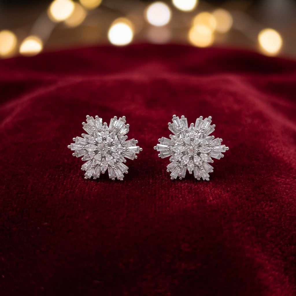 A pair of snowflake-shaped stud earrings set with clear crystals on a deep red velvet surface with warm bokeh lights in the background.