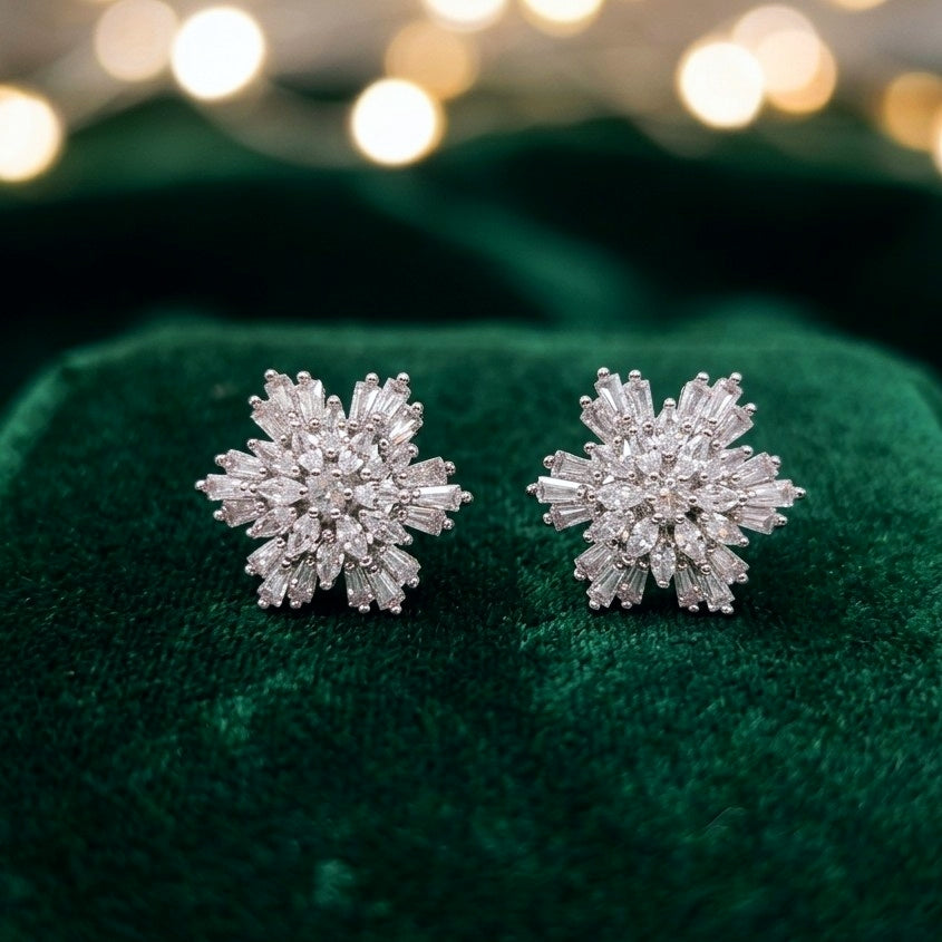 A pair of snowflake-shaped stud earrings with clear baguette and round stones, displayed on a dark green velvet surface with a blurred festive bokeh background.