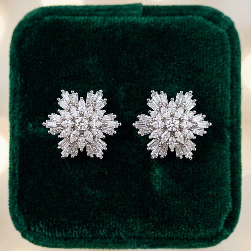 A pair of snowflake-shaped stud earrings made of sparkling clear stones in a silver-toned setting, displayed on a dark green velvet jewelry box against a soft, blurred background of warm bokeh lights.