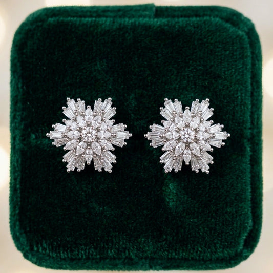 A pair of snowflake-shaped stud earrings made of sparkling clear stones in a silver-toned setting, displayed on a dark green velvet jewelry box against a soft, blurred background of warm bokeh lights.