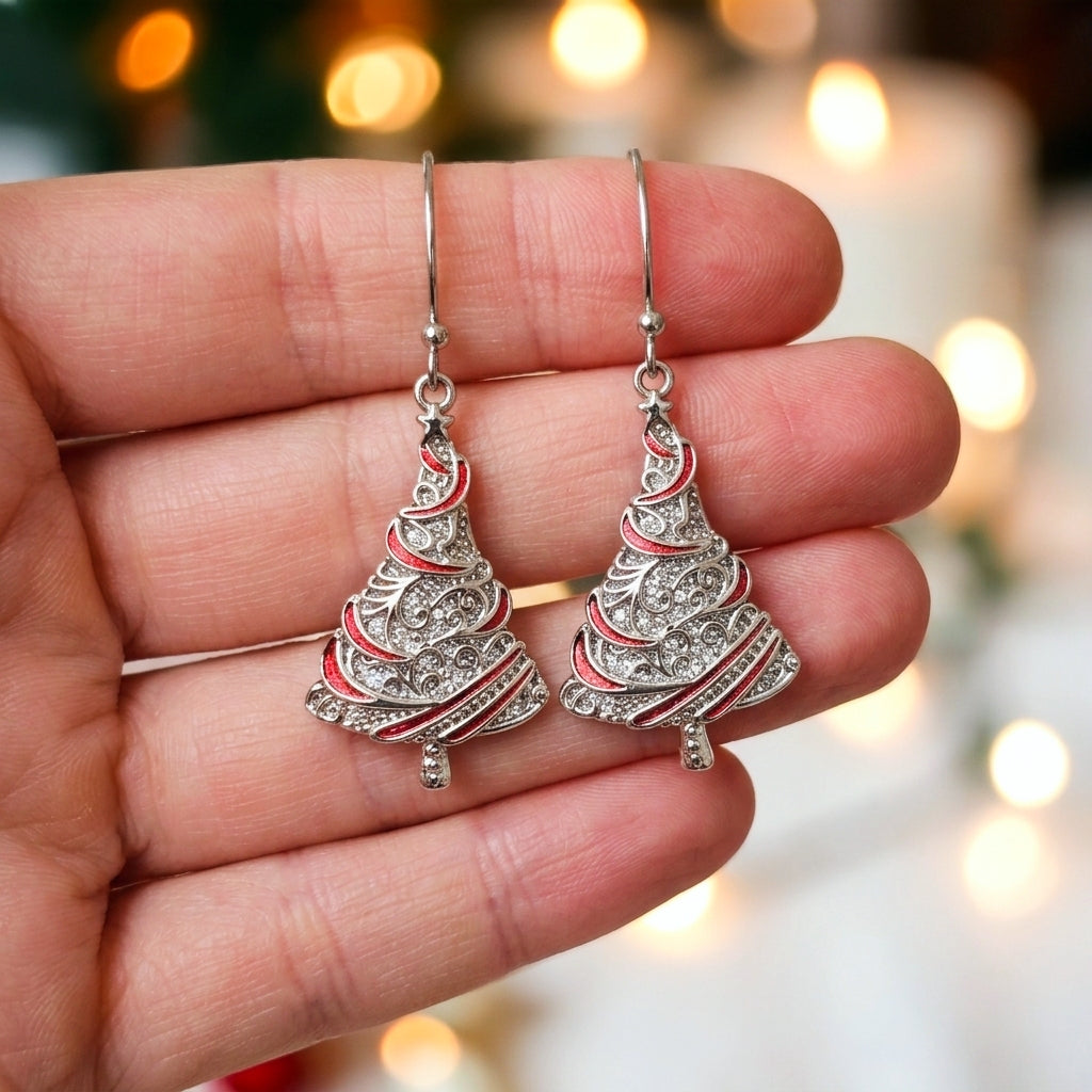 A pair of silver Christmas tree dangle earrings with red enamel accents and clear rhinestones, held in a person's hand against a warm, blurred holiday background.