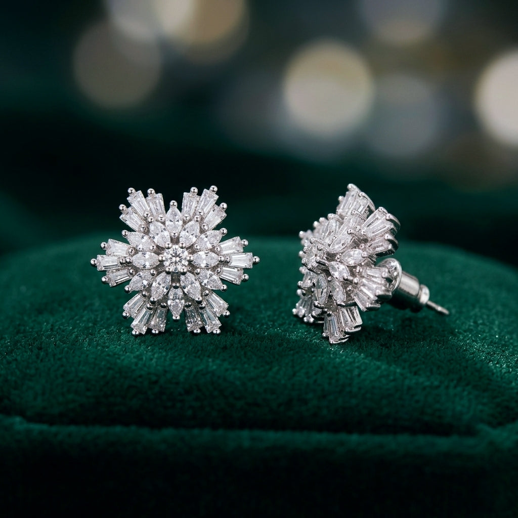 A pair of silver-toned snowflake stud earrings with baguette-cut clear stones, displayed on a dark green velvet jewelry box against a soft, blurred bokeh background.