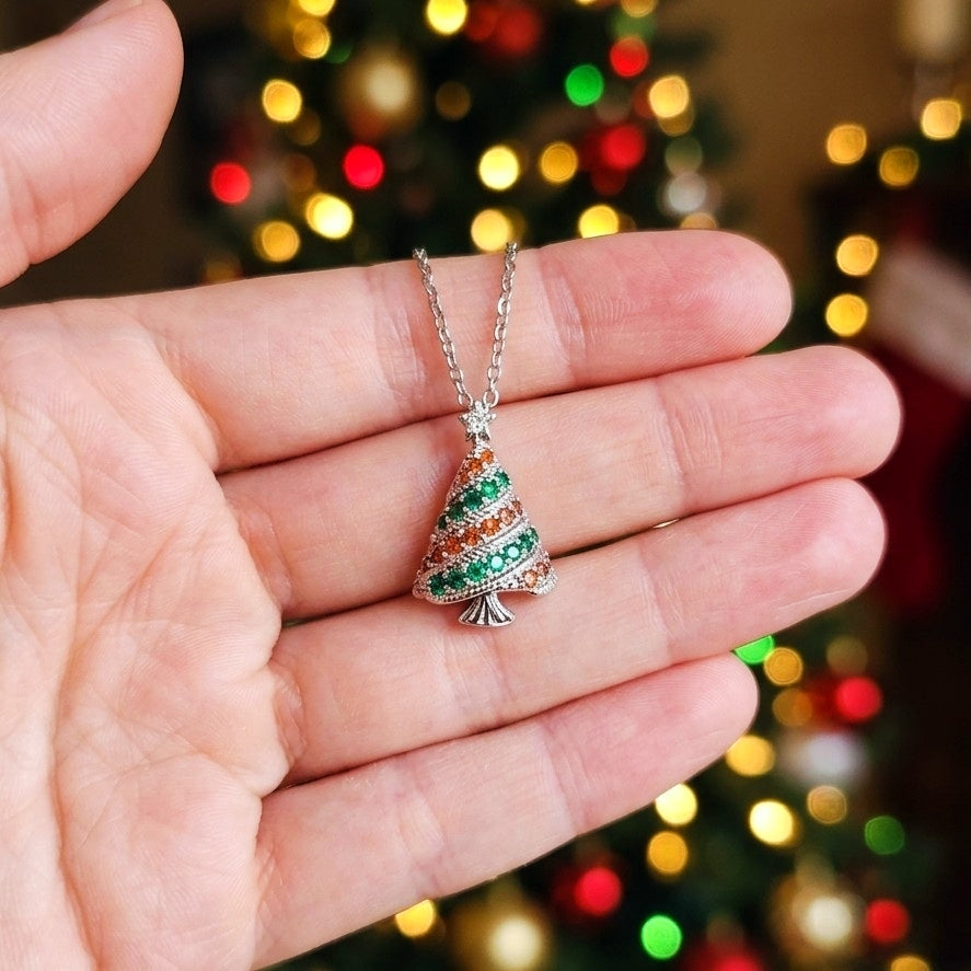 A silver Christmas tree pendant necklace with green and orange cubic zirconia crystals, held in a person's hand against a blurred background of a lit Christmas tree.