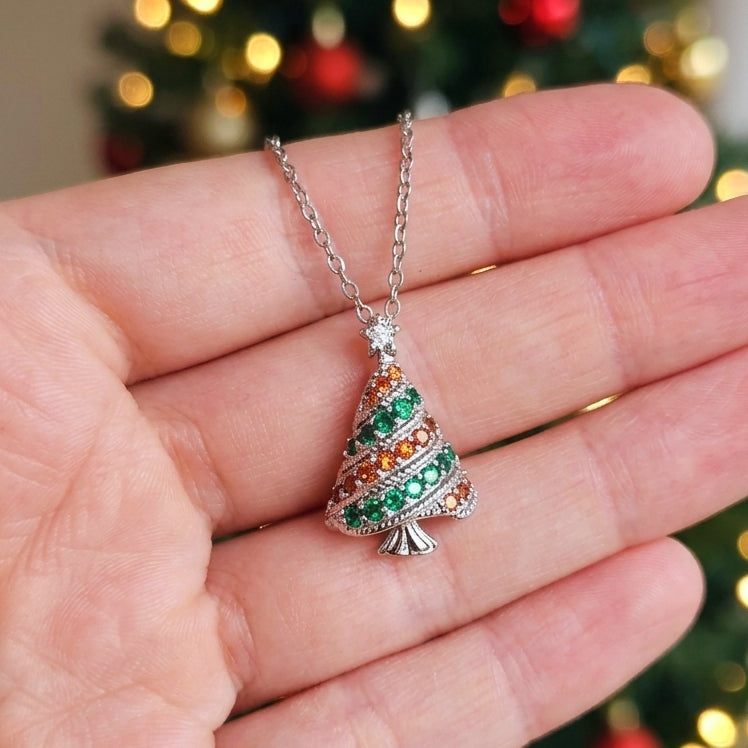 A silver Christmas tree pendant necklace with green and orange cubic zirconia crystals, held in a person's hand against a blurred background of a lit Christmas tree.