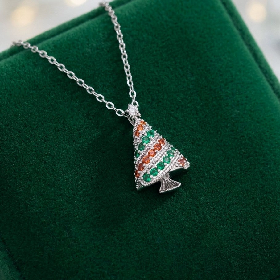 A silver Christmas tree pendant necklace with green and orange cubic zirconia crystals, displayed on a dark green velvet jewelry box.
