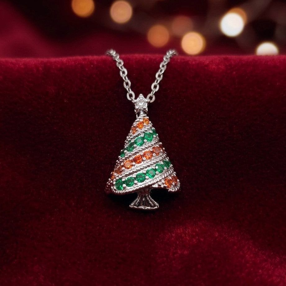A silver Christmas tree pendant necklace adorned with green and orange cubic zirconia crystals, resting on a deep red velvet surface with a warm bokeh background.