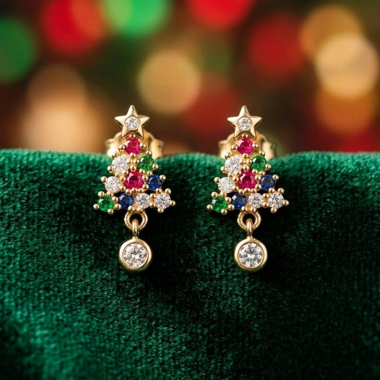 A pair of gold Christmas tree stud earrings with colorful gemstones and clear crystal drops, displayed on a textured dark green velvet surface with a blurred festive bokeh background.