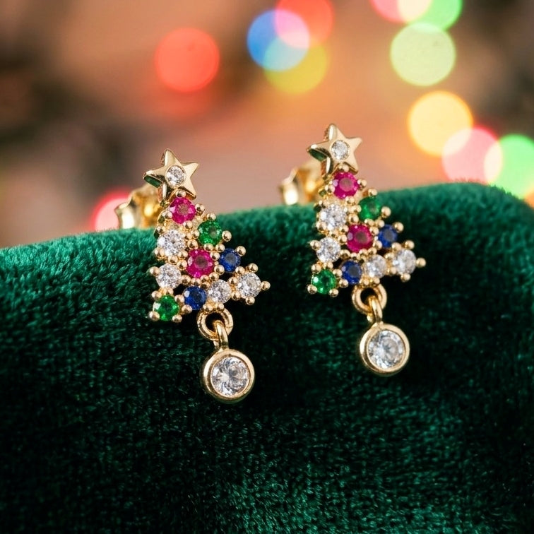 A pair of gold dangle earrings shaped like Christmas trees, embellished with clear cubic zirconia stones and colored gemstones in pink, blue, and green, displayed on a dark green velvet surface with a blurred festive bokeh background.