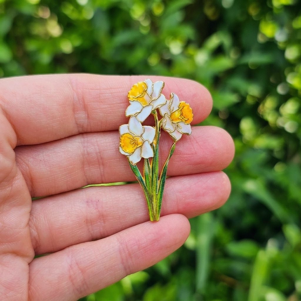 A small enamel daffodil flower brooch with white petals, yellow centers, and green stems is held on a person's fingers against a blurred green outdoor background.