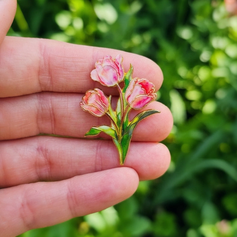 A close-up of a hand holding a small enamel pin shaped like three pink tulips with green leaves, set against a blurred green outdoor background.
