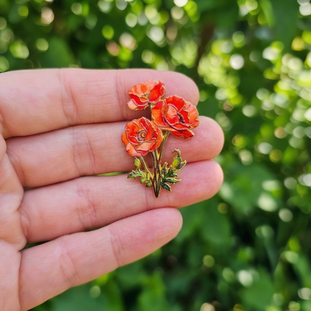 A small golden floral enamel pin with three poppy flowers and green leaves held on a person's fingers against a blurred green outdoor background.