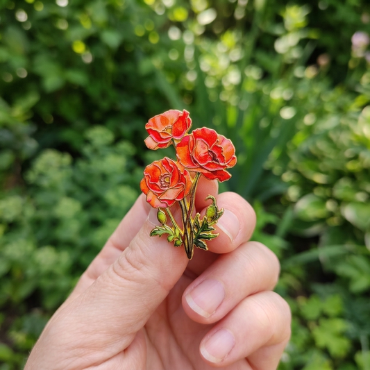 A hand holding a small enamel pin shaped like three red poppy flowers with green leaves, set against a blurred green garden background.
