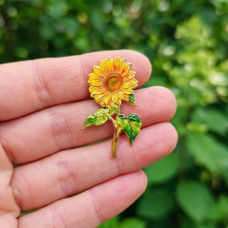 A small enamel sunflower pin held on an open hand, with a blurred green outdoor background.
