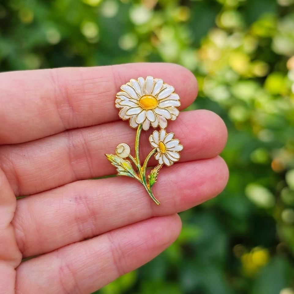A small daisy flower brooch with white petals, yellow centers, and green stems is displayed on a person's hand against a blurred green outdoor background.