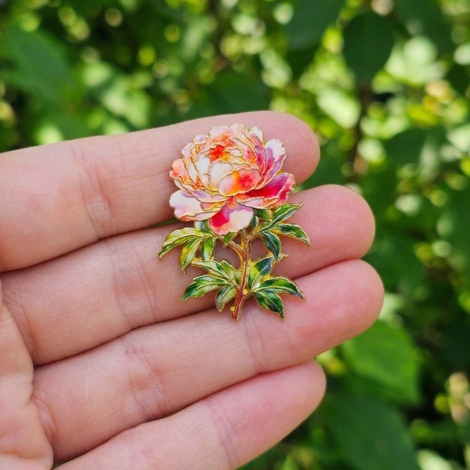 A close-up of a hand holding a small enamel peony flower pin with pink, orange, and white petals and green leaves against a blurred green outdoor background.