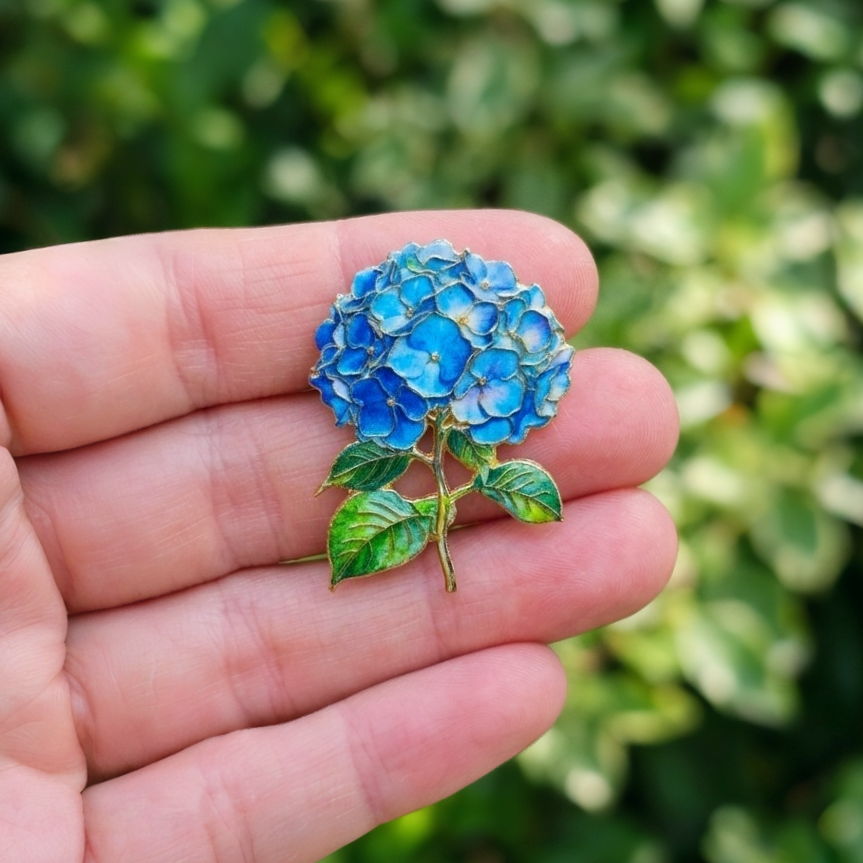 A close-up of a hand holding a detailed blue hydrangea flower brooch with green leaves against a blurred green outdoor background.