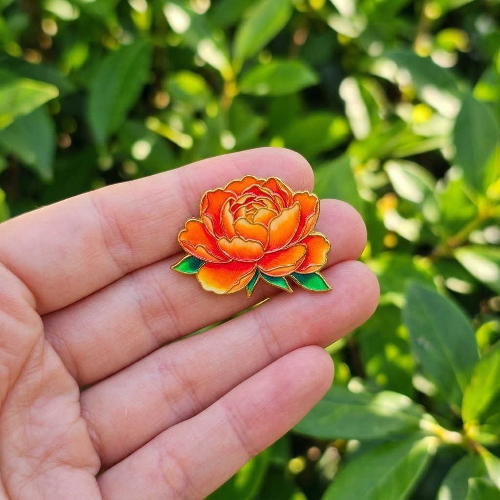 A small enamel peony flower pin with orange and yellow petals and green leaves, held on a person's finger against a blurred green outdoor background.