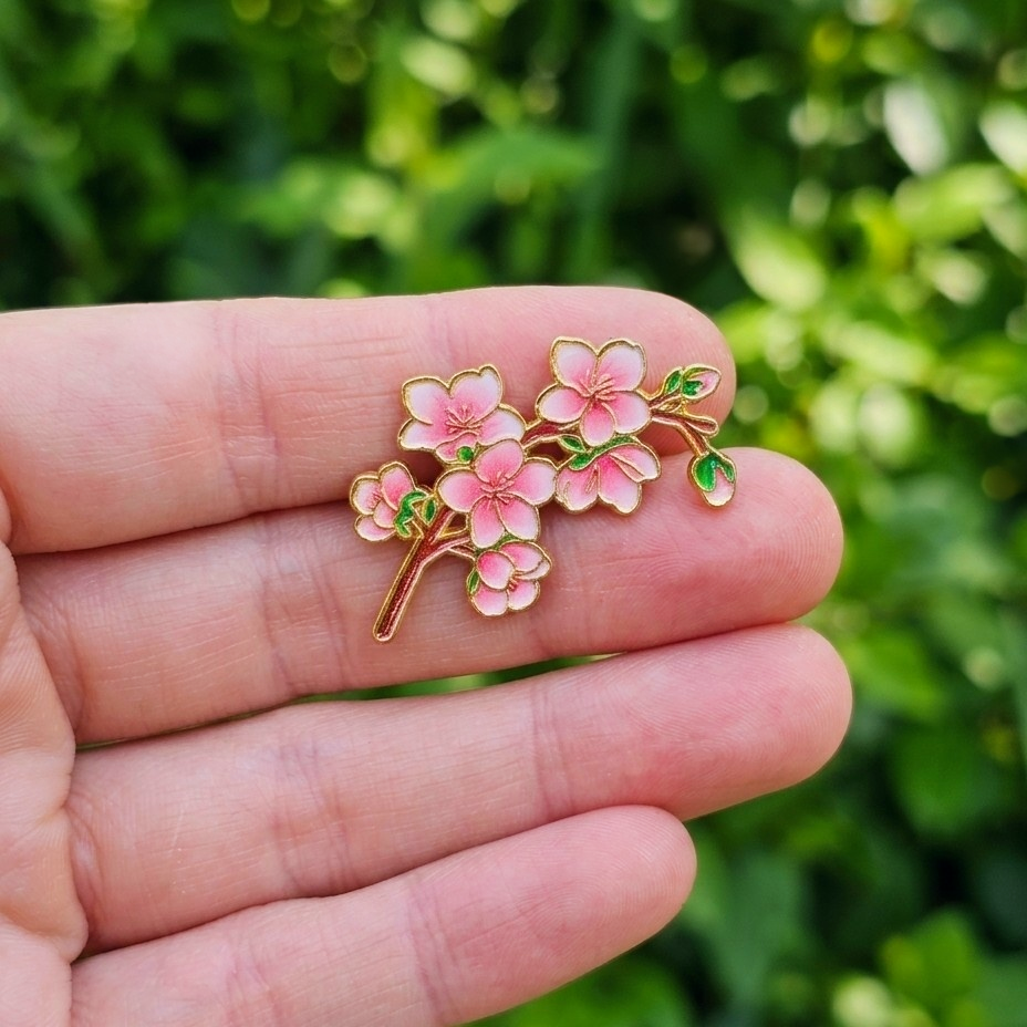 A small enamel pin shaped like pink cherry blossoms on a branch, held on a person's fingers against a blurred green outdoor background.