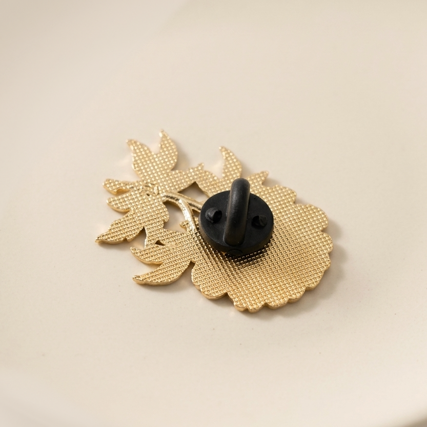 The back of a gold metal pin with a black rubber fastener, photographed on a plain light beige background.