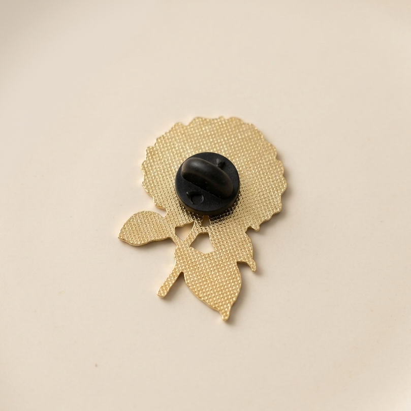 The back of a gold metal flower-shaped pin with a black plastic clutch fastener in the center, shown on a plain off-white background.