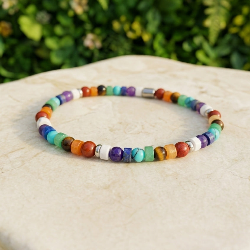 A colorful beaded bracelet made of small round and square gemstone-like beads in multiple hues, displayed on a light beige stone surface with a blurred green garden background.
