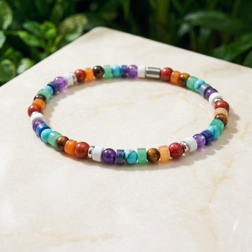 A colorful beaded stretch bracelet made of round and square gemstone beads in multiple vibrant colors, displayed on a light beige stone surface with a blurred green plant background.