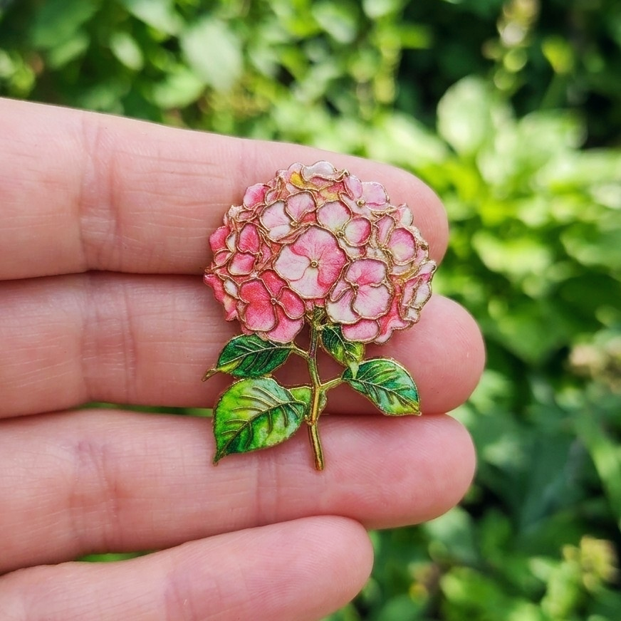 A close-up of a hand holding a small enamel pink hydrangea flower pin brooch with green leaves and gold-tone outlines, set against a blurred green outdoor background.