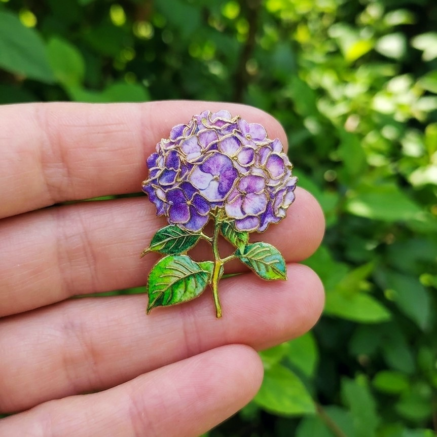A close-up of a hand holding a detailed purple hydrangea flower pin brooch with green leaves and gold metal outlines, set against a blurred green outdoor background.