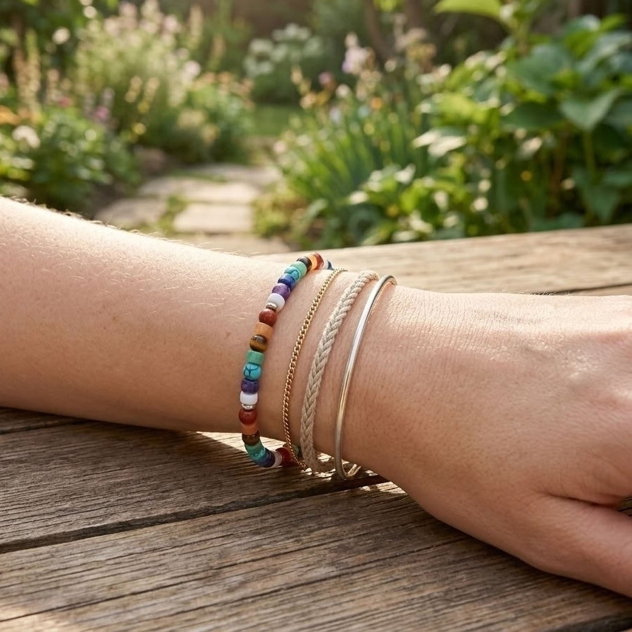 A close-up of a person's wrist wearing multiple bracelets, including a colorful beaded bracelet, a braided cord bracelet, a thin gold chain, and a silver bangle, resting on a wooden table with a blurred garden background.