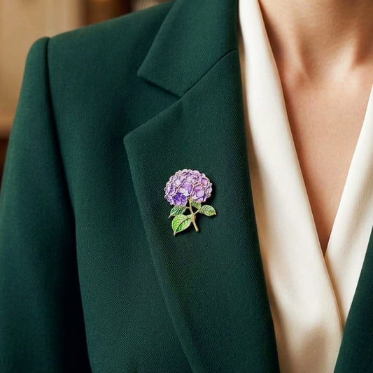 A close-up of a person wearing a dark green blazer over a cream satin V-neck top, featuring a small purple and green enamel hydrangea brooch pinned to the lapel.