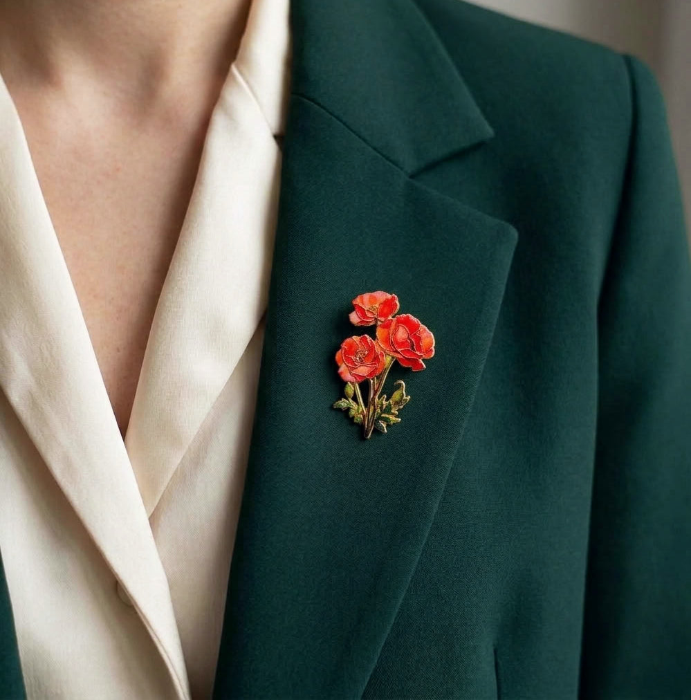 A close-up of a person wearing a dark green blazer over a cream satin blouse, featuring a small red poppy flower brooch pinned to the lapel.