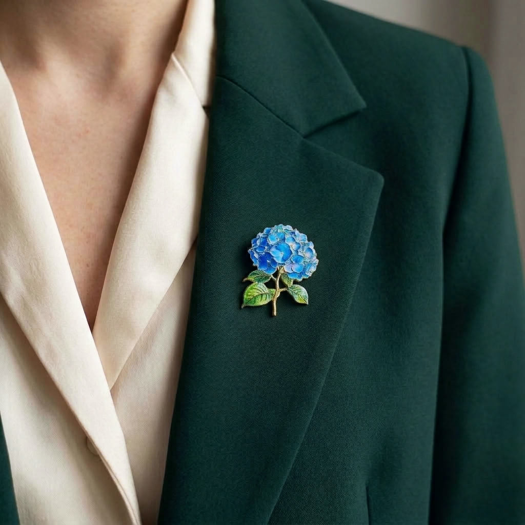 A close-up of a person wearing a dark green blazer over a cream satin blouse, featuring a blue and green enamel hydrangea brooch pinned to the lapel.