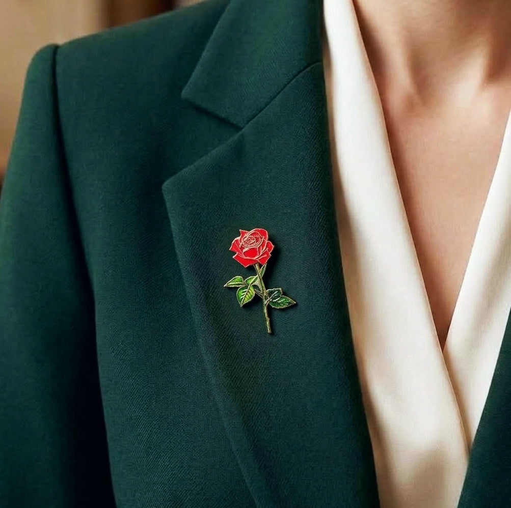 A close-up of a dark green blazer worn with a cream satin V-neck blouse, featuring a small red rose lapel pin on the left lapel.