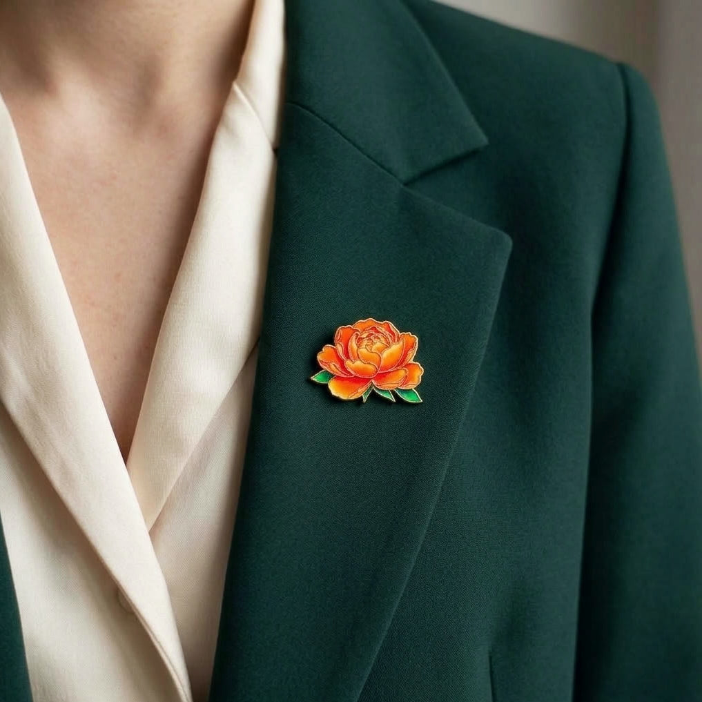 A close-up of a person wearing a dark green blazer over a cream satin blouse, featuring a small orange peony brooch with green leaves on the blazer chest.