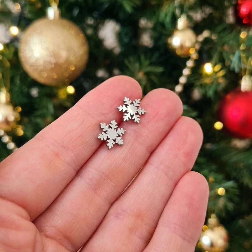 A hand holding small snowflake stud earrings made from stainless steel with a Christmas tree in the background.