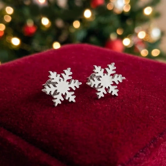 A pair of silver snowflake stud earrings resting on a deep red velvet jewelry box, with a blurred Christmas tree in the background.