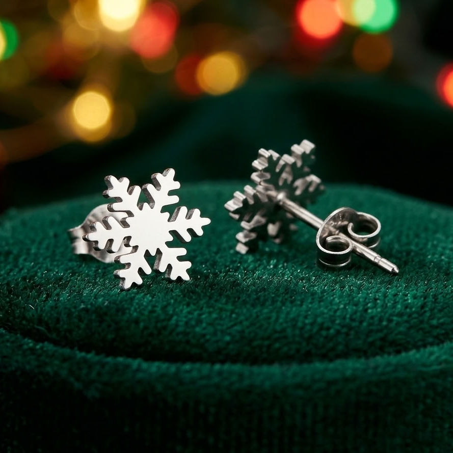 A pair of silver snowflake stud earrings displayed on a textured dark green velvet surface with a blurred festive background of warm holiday lights.