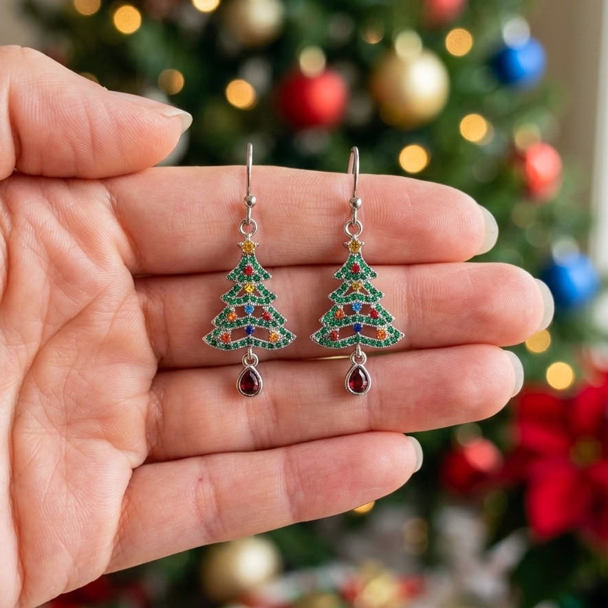 A pair of silver Christmas tree drop earrings with colorful cubic zirconia crystals and red teardrop charms, held in a hand with a blurred Christmas tree in the background.