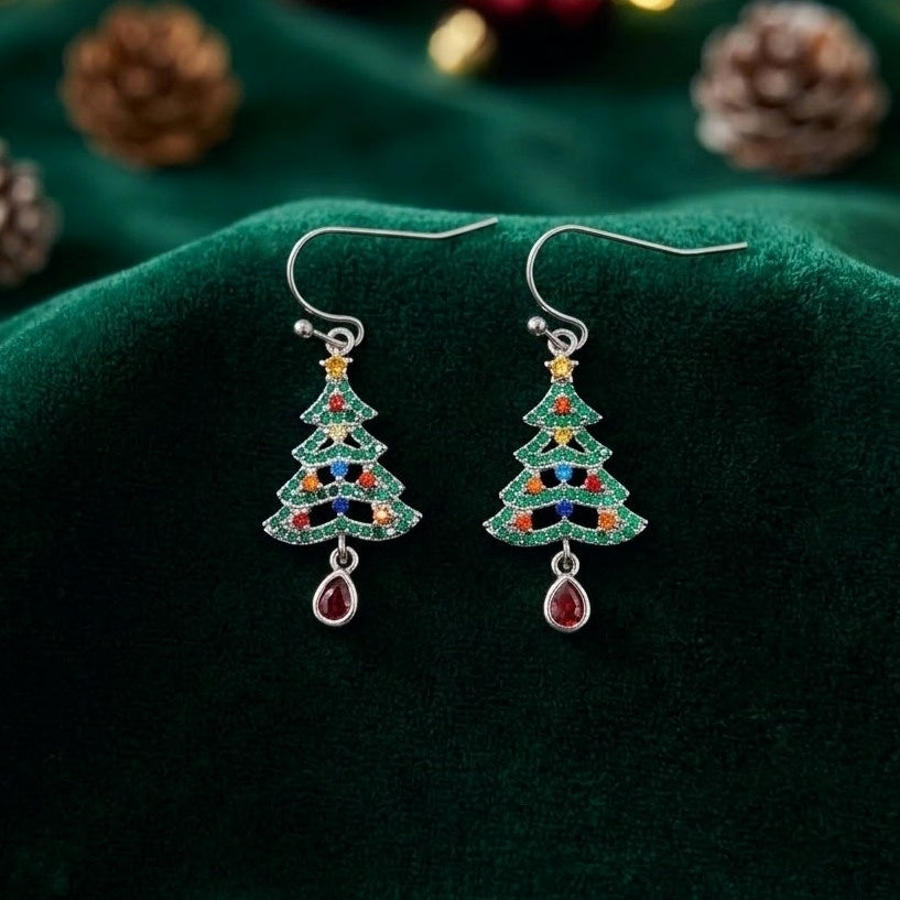 A pair of silver-toned dangle earrings shaped like Christmas trees, decorated with green, red, blue, and yellow cubic zirconia crystals, and each hanging from a teardrop-shaped red gem, displayed on a dark green velvet fabric background with pinecones.