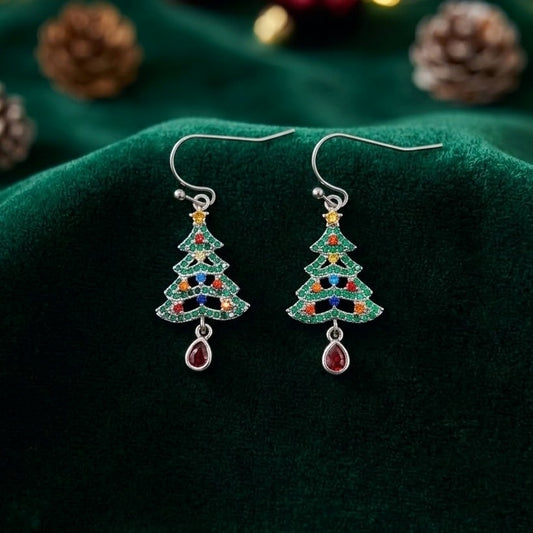 A pair of silver-toned dangle earrings shaped like Christmas trees, decorated with green, red, blue, and yellow cubic zirconia crystals, and each hanging from a teardrop-shaped red gem, displayed on a dark green velvet fabric background with pinecones.