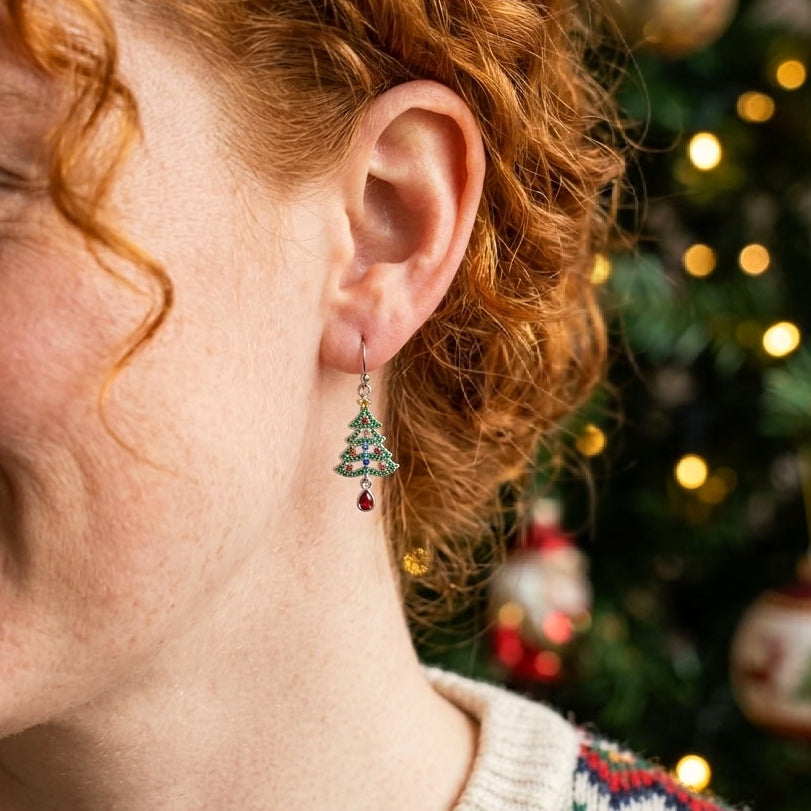 A close-up of a person with red hair wearing a dangling Christmas tree earring with green cubic zirconia crystals and a red teardrop charm, set against a blurred Christmas tree background with warm lights.