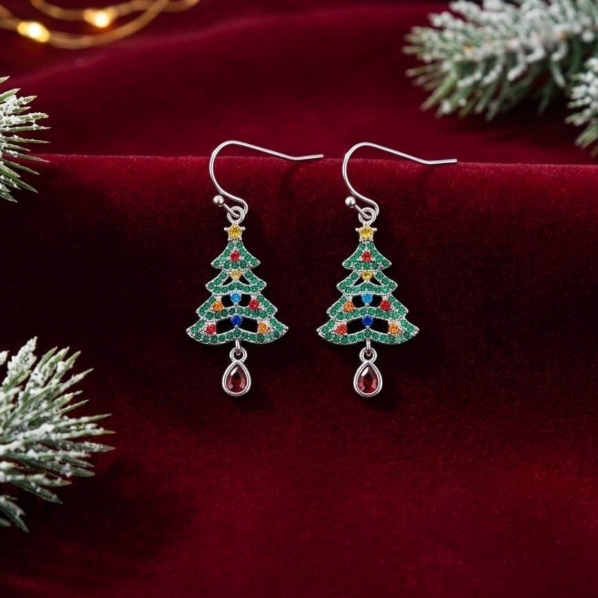 A pair of silver dangle earrings shaped like Christmas trees, decorated with green, red, and blue cubic zirconia crystals and a dangling pink teardrop gem, displayed on a dark red velvet surface with festive pine branches.