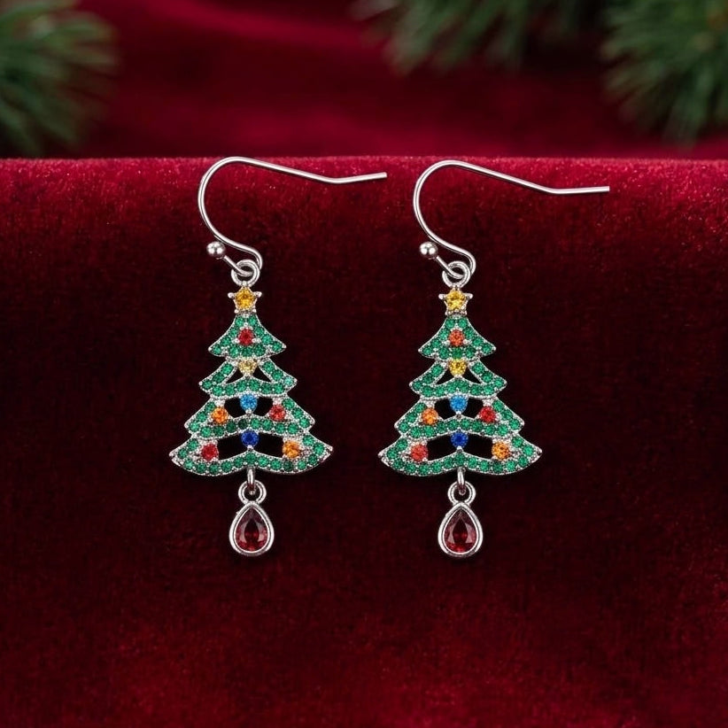 A pair of silver-toned dangle earrings shaped like Christmas trees, decorated with green cubic zirconia crystals and small multicolored accents, displayed on a deep red velvet surface with blurred green pine branches in the background.