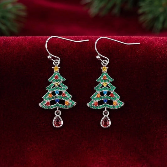 A pair of silver-toned dangle earrings shaped like Christmas trees, decorated with green cubic zirconia crystals and small multicolored accents, displayed on a deep red velvet surface with blurred green pine branches in the background.